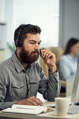 Serious young bearded call center operator in headset with microphone sitting at desk and using laptop