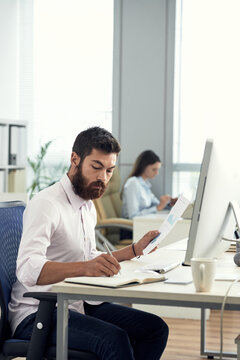 Serious busy young bearded accountant sitting at desk with computer and working with graph in office