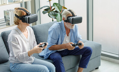 Caucasian old senior elderly grandparents couple wears virtual reality goggles headset. gray bearded and hair grandpa husband and grandma wife holding joysticks playing vr videogame in living room