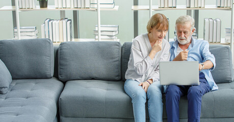 Caucasian old senior elderly grandparents lover couple in casual wear sitting together on couch in living room at home looking to laptop computer screen learning new lesson shopping online from store