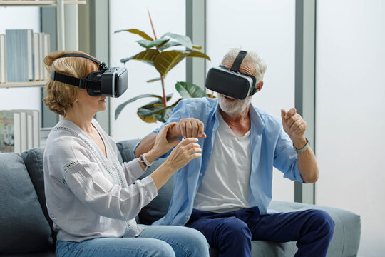 Caucasian old senior elderly grandparents couple wears virtual reality goggles headset. gray bearded and hair grandpa husband and grandma wife sitting together playing vr videogame in living room