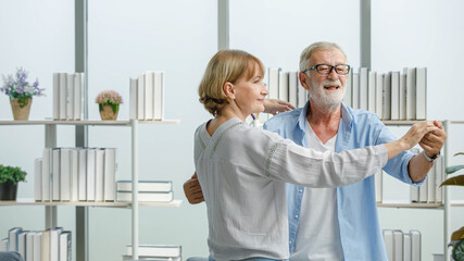 Obraz premium Caucasian old senior elderly happy grandparents lover married couple standing smiling look at each other face listening to slow waltz music dancing together in living room at home in front bookshelf