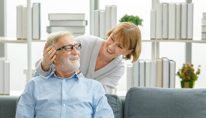 Caucasian old senior elderly happy supportive lovely female grandma wife helping massaging grey bearded hair husband head sitting smiling on cozy sofa to relief pain and stress in living room at home