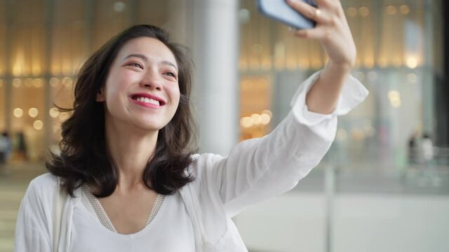 happy charming young asian woman walking in the city shopping mall holding mobile phone video chatting with friend online slow motion 4k clip of urban people lifestyle