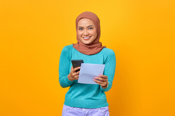 Happy young Asian woman holding smartphone and bill isolated on yellow background