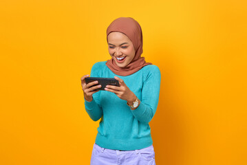 Cheerful young Asian woman playing a video game on a smartphone on yellow background