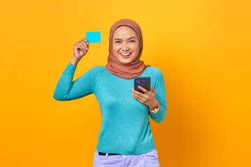 Smiling young Asian woman showing credit card and holding mobile phone on yellow background