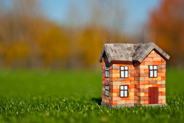 Brick toy house on the grass. Orange house on a green meadow. The concept of own housing and construction. Yellow trees in the background.