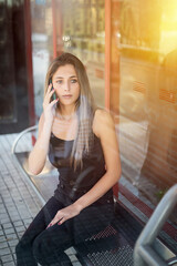 Young blonde girl talking on the phone waiting seated at a bus stop