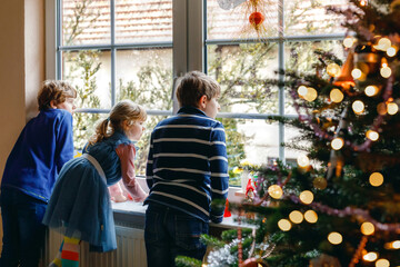 Fototapeta na wymiar Three cute children sitting by window on Christmas eve. Two school kid boys and toddler girl, siblings looking outdoor and dreaming. Family happiness on traditional holiday
