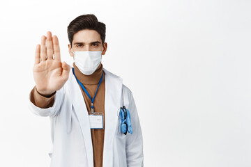 Covid-19, quarantine and healthcare concept. Serious doctor in robe and face mask, showing stop sign, extend one palm in disapproval, reject smth, standing over white background