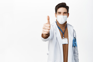 Coronavirus and vaccination concept. Young doctor in medical face mask and robe, showing thumb up, approve, like smth good, standing over white background