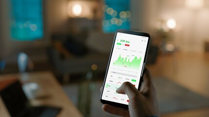 Close-up Shot of Smartphone. Man is Using Stock Market App to Check Share Prices. Graph is Going Up Making a Great Profit. In the Background Cozy Homely Atmosphere, Dusk Time.