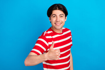 Photo of funky charming young man wear striped t-shirt smiling showing thumb up isolated blue color background