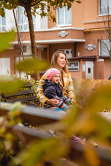 Mom and daughter are sitting on city bench during autumn walk. Fall colors