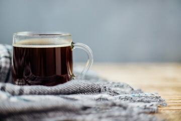 A warm plaid scarf with fringe lies on a light wooden background. A glass mug with black coffee stands on a soft cloth.