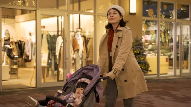 happy asian mom pushing a baby carriage and doing window shopping for Christmas presents on downtown street with beautiful light decoration in the evening.