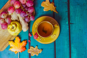 Beautiful fall still life with yellow cup on saucer. Bunch of red grapes, ripe agave, ginger root lie on cutting board. Fallen oak, maple leaves. Wooden green emerald background with space for text.
