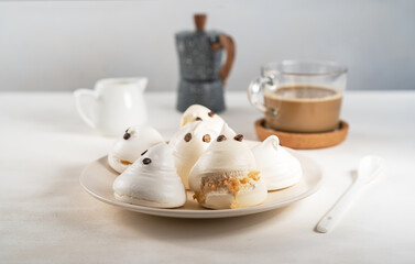 Fototapeta na wymiar Meringue in a form of ghosts on the plate with cup of coffee, milk jar and coffe pot on the light background