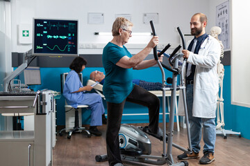 Aged woman using electrical bicycle for physical exercise to recover from injury. Senior patient doing gymnastics with stationary bike while doctor giving support for physiotherapy