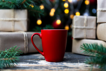 Coffee in a red cup on the rustic table with wrapped gifts, Christmas tree lights and spruce branches in the background