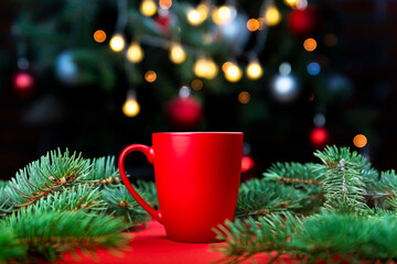 Morning coffee in a red cup on the red surface with Christmas tree lights and spruce branches in the background