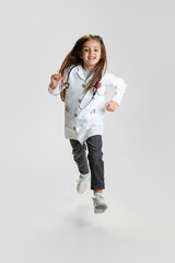 Full-length portrait of cute beautiful little girl, child in image of doctor wearing white lab coat running isolated on white studio background