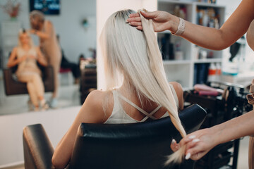 Hairdresser female making hair extensions to young woman with blonde hair in beauty salon....
