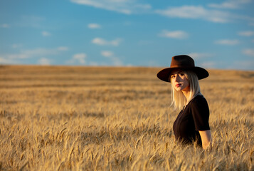 Naklejka na ściany i meble Blonde woman in black dress and hat with scarf in wheat field in autumn season time