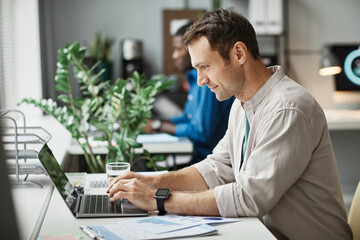 Side view portrait of smiling adult man using laptop while enjoying work in office, copy space - obrazy, fototapety, plakaty