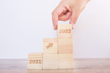 Hand puts wooden cube, arrow upwards on wooden blocks from 2021 to 2022 on white background.