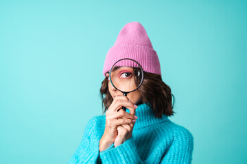 Cozy portrait of a young woman in a knitted blue sweater and a pink hat with bright makeup holding a magnifying glass, fooling around, having fun - obrazy, fototapety, plakaty