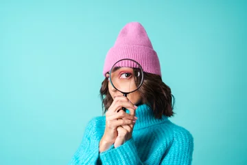 Foto op Aluminium Cozy portrait of a young woman in a knitted blue sweater and a pink hat with bright makeup holding a magnifying glass, fooling around, having fun © Анастасия Каргаполов