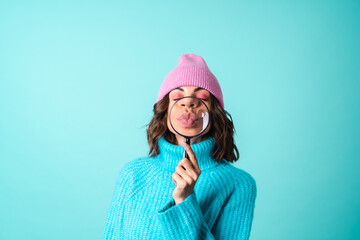 Cozy portrait of a young woman in a knitted blue sweater and a pink hat with bright makeup holding a magnifying glass, fooling around, having fun