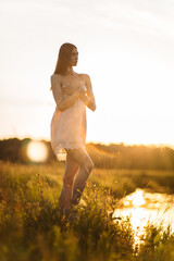 a young beautiful girl, of model appearance, in a shirt, stands among the landscape on the river bank, the setting sun illuminates her figure