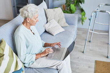 Side view portrait of white haired senior woman using laptop while relaxing on sofa at home, copy space