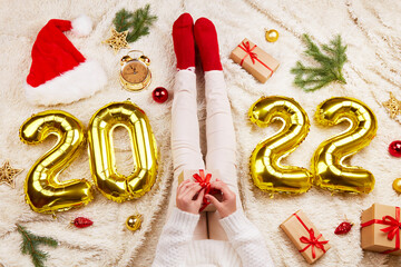 2022 Happy New Year greeting card. The girl is holding a gift in her hands, next to toys and Christmas decorations, gold foil balloons with the numbers 2022. 