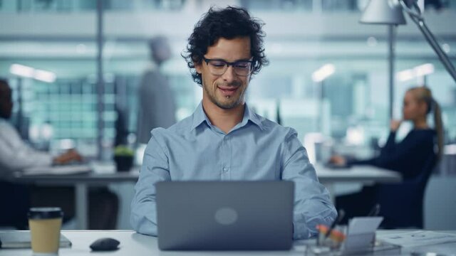 Modern Office Businessman Working on Computer, Smiling, Looking at Camera. Portrait of Successful Latin IT Software Engineer Working on a Laptop at his Desk. Front View Static Shot