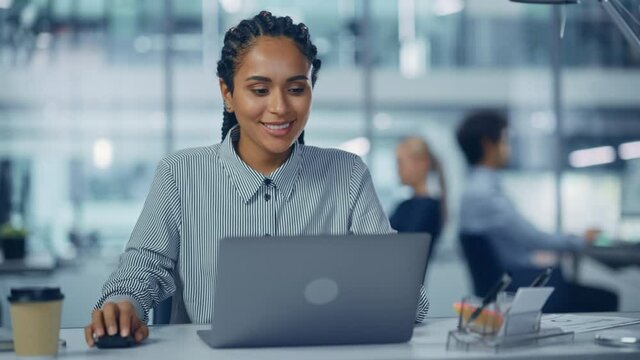 Multi-Ethnic Office: Professional Black Female IT Engineer Working on Desktop on Laptop Computer. Successful, Smiling Businesswoman with Braided Hair Does Creative Job. Confident Product Designer