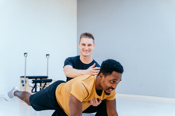 Happy smiling male coach or instructor help black man client at individual pilates class in fitness studio. Multiethnic man have stretching pilates session with personal trainer. Sports concept.