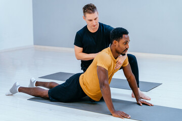 Handsome male pilates trainer helps to african man, individual training participant, do right...
