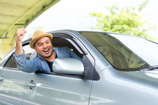 Happy male driver smiling while sitting in a car with open front window. Young asian man smile and looking through window enjoying trip. Man driving his car to travel on his holiday vacation time.