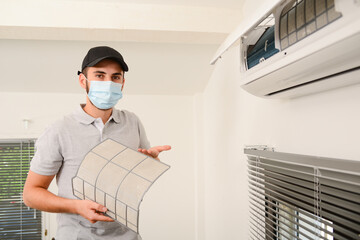 Fototapeta na wymiar handsome young man electrician with surgical mask cleaning air filter on an indoor unit of air conditioning system in client house