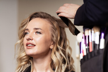 Unrecognizable hair master makes curls with curling iron for young woman with blond hair. Portrait of happy blonde model.