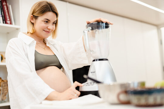 White pregnant woman smiling and making smoothy at home
