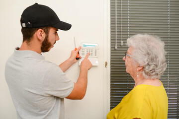handsome young man installing house security anti burglary camera and siren alarm in a senior woman home - obrazy, fototapety, plakaty