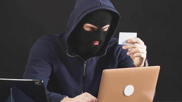 Man hacking laptop system, want to hack bank card, sit in black mask, black background