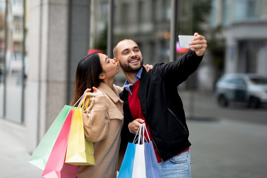 Loving interracial couple with colorful bags taking selfie on mobile phone in front of shopping mall