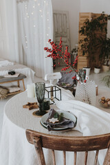 Elegant aesthetic Christmas dinner concept. Table with grey linen table cloth, red berries branch bouquet, gingerbread cookies, glass, plates. Christmas celebration evening