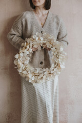 Beautiful young woman in beige woolen sweater and silk skirt hold Christmas wreath made of dried gold lunaria leaves in front of neutral dusty pink wall. Elegant aesthetic fashion Christmas background
